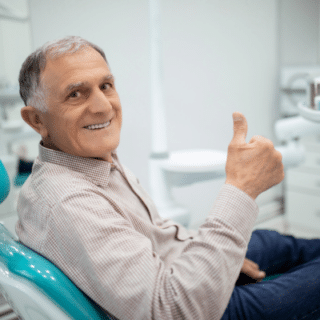Older man sitting in a dental chair giving the thumbs up while smiling, representing senior dental health