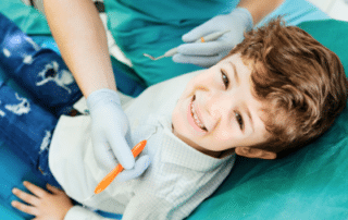 a boy sitting in a chair with a dentist holding an orange tool, representing kids dental hygiene