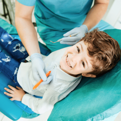a boy sitting in a chair with a dentist holding an orange tool, representing kids dental hygiene